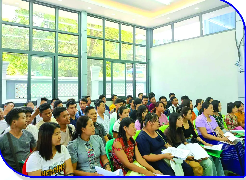 A classroom filled with seated candidates holding papers, tall windows along one wall