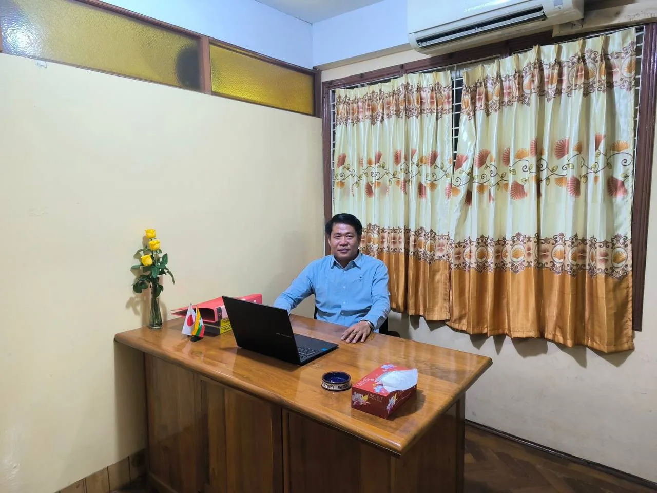 Min Naing Win seated at his desk in the company office, with Myanmar and Japanese desk flags beside a laptop