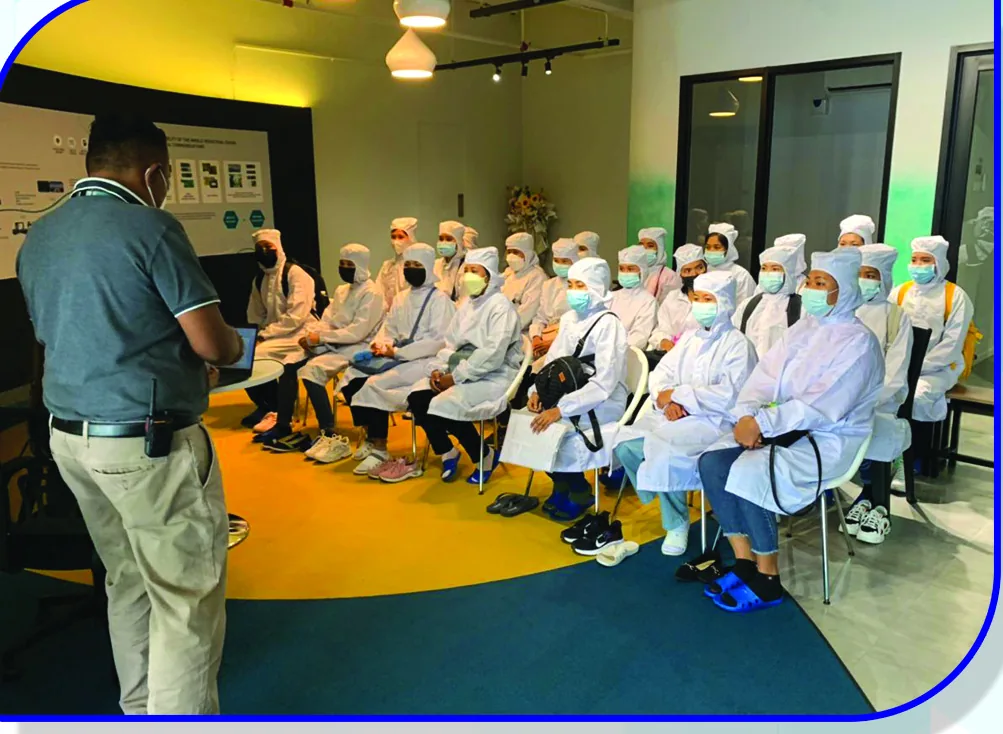An instructor addressing a seated group wearing protective gowns, caps and masks